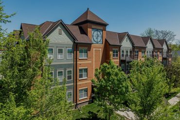 Apartment building with a clock tower surrounded by green trees under clear blue sky.