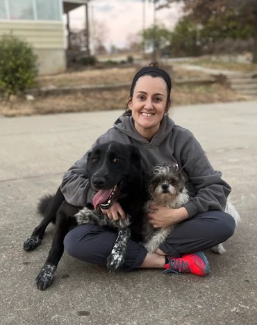 Woman sitting on pavement hugging two dogs, smiling warmly.