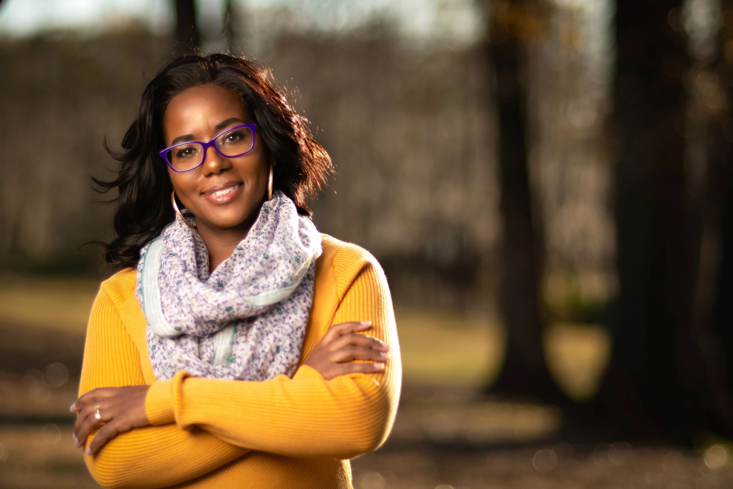 Black woman, flowing hair wearing purple framed glasses; wearing a yellow sweater with folded arms.