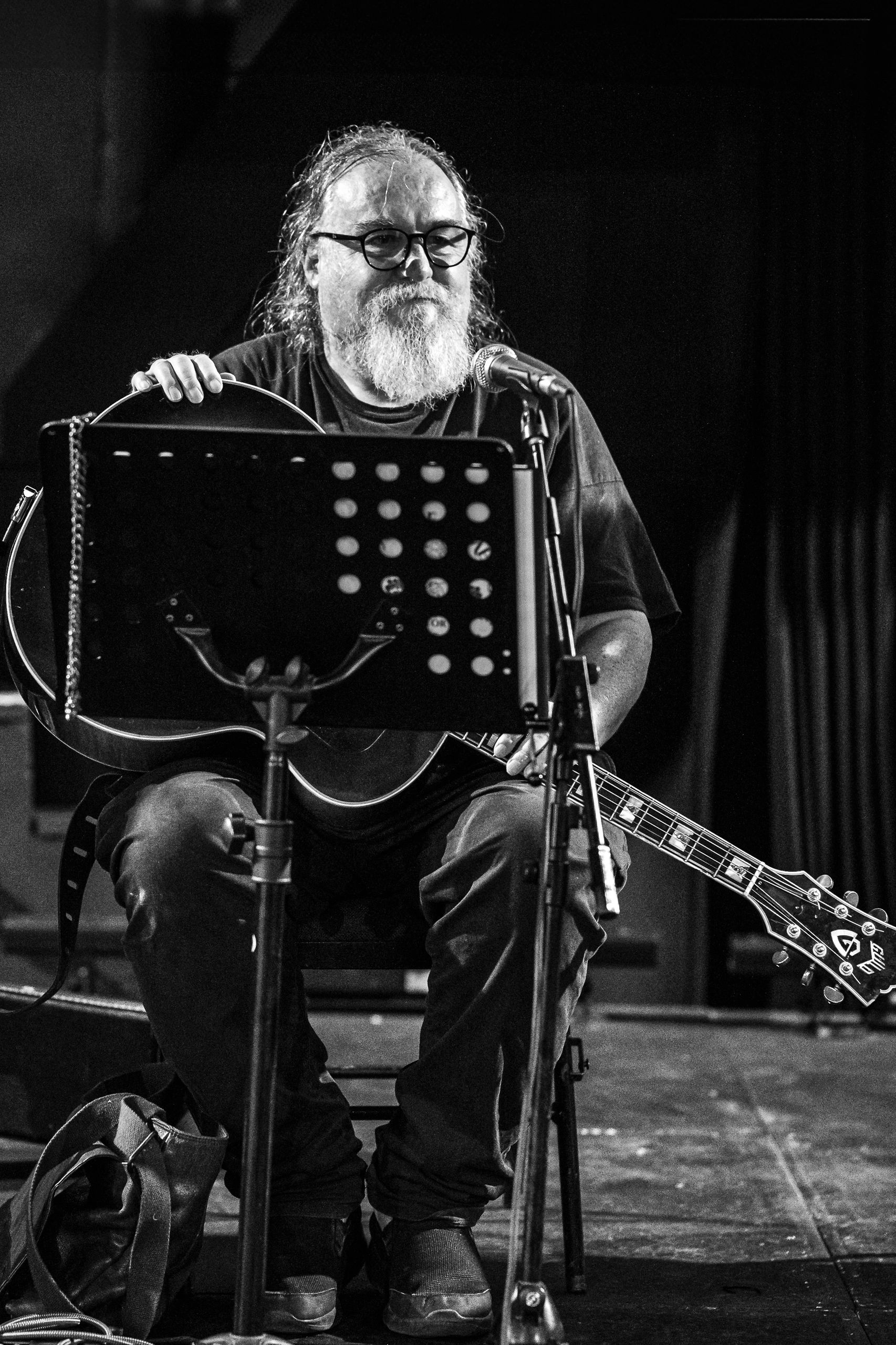 Bearded man with glasses playing guitar on stage.