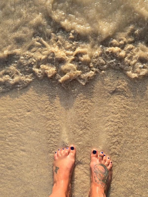 Feet with tattoos and navy blue nail polish at sandy beach shore.