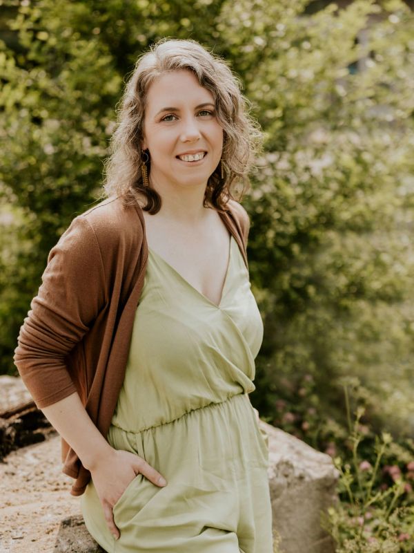 Woman in a light green dress and brown cardigan smiling outdoors.