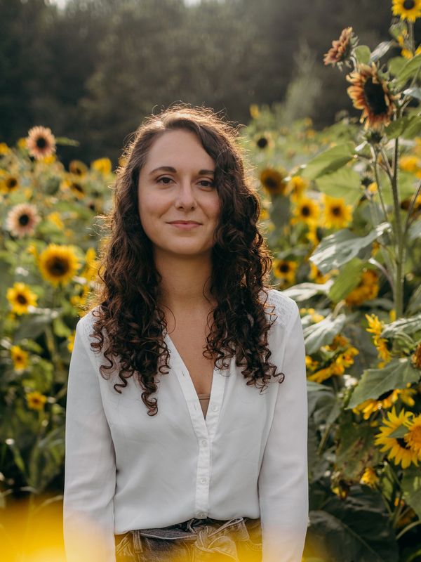 Woman with curly hair stands among vibrant sunflowers, wearing a white blouse.