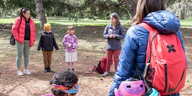 Adults and children gathered outdoors in a wooded area, engaged in conversation.