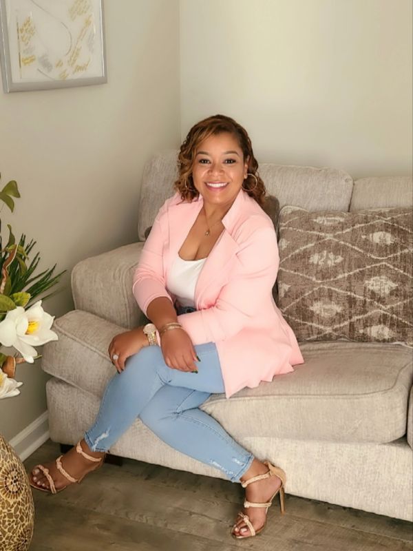 Smiling woman sitting on a beige couch in a pink blazer and light blue jeans.