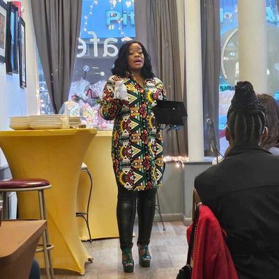 A woman in colorful dress speaks to a seated audience in a cozy room.