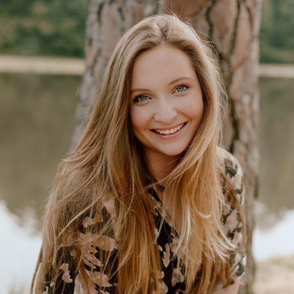 Smiling woman with long blonde hair outdoors near a tree and water.