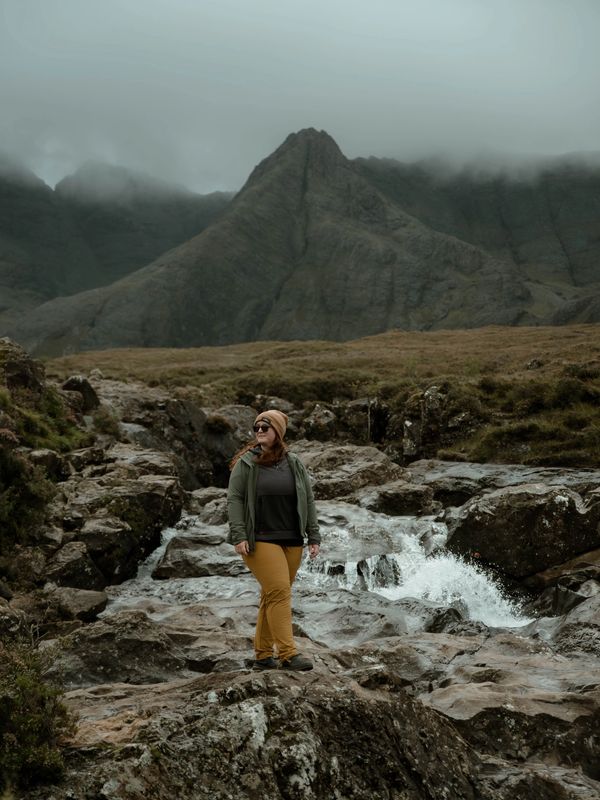 Woman hiking near a rocky stream with misty mountains in the background.
