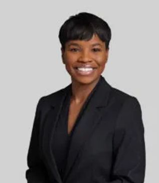 Professional woman in a black blazer smiling confidently against a gray background.