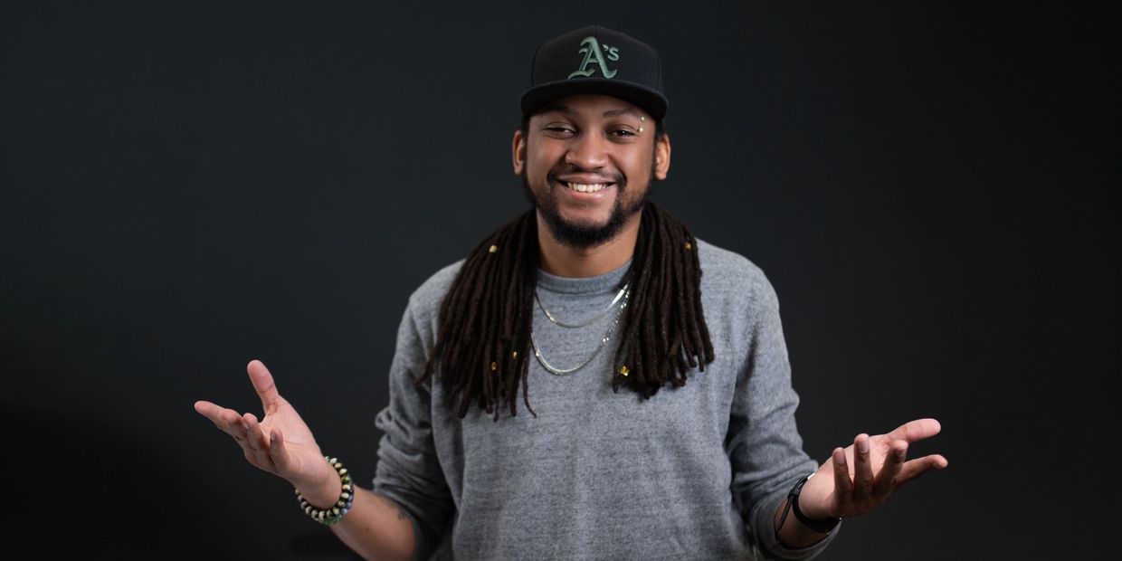 Smiling man with dreadlocks wearing a gray sweater and black cap gestures with open hands.