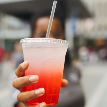 Hand holding a cold, red and clear gradient drink with a straw in a plastic cup.