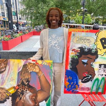 A smiling woman stands outdoors with two colorful paintings.