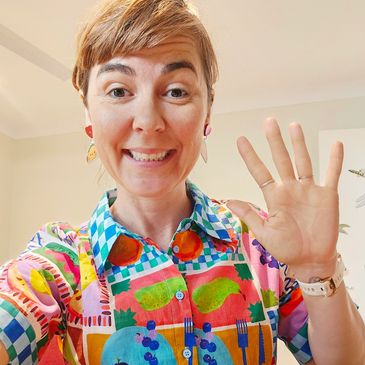 Smiling woman waving, wearing a colorful patterned shirt indoors.