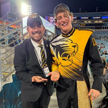 Two smiling people at a stadium holding hearing aids, one in a suit and one in a band uniform.
