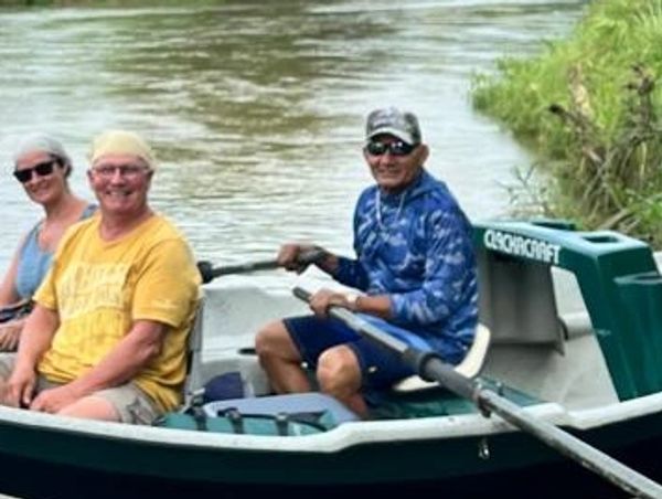 Three people smiling in a small boat on a river near grassy banks.