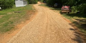 A gravel road winds through a green, wooded rural area under clear blue skies.
