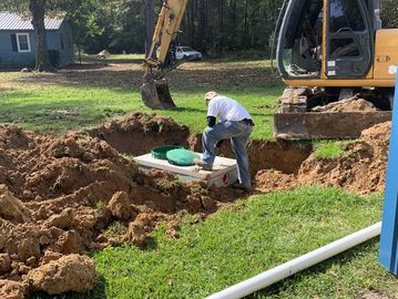 A worker inspects a septic tank installation with an excavator nearby.