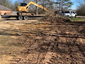 Excavator on trailer and white truck near a pile of dirt and branches in a yard.