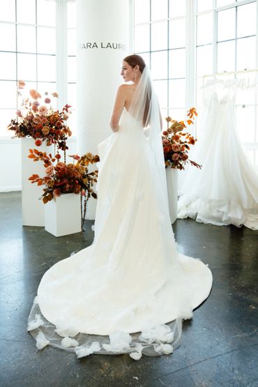 Bride modeling an elegant white wedding dress with floral details and a long veil indoors.