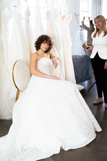 A woman in a white wedding dress sits elegantly amid other dresses.
