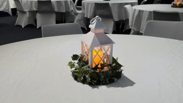 White lantern with a glowing candle and green leaves on a round table.