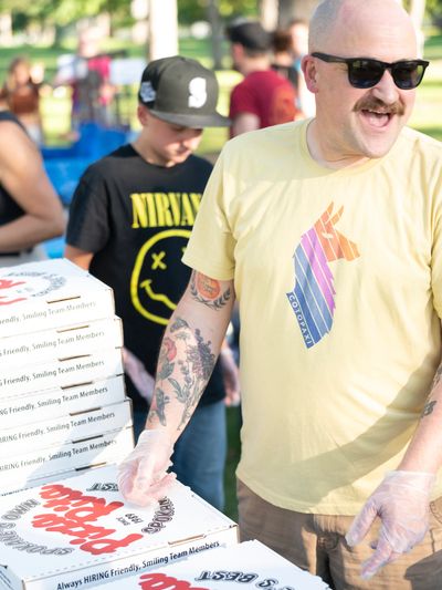 Man with tattoos and sunglasses serving pizza at an outdoor event.