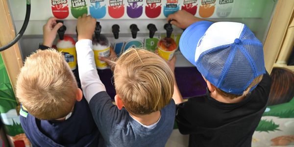 Three kids getting slushies from a machine with various flavors.
