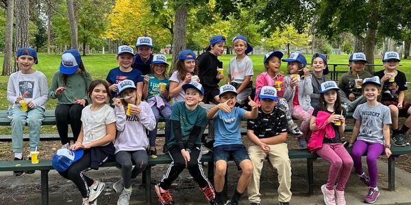 A group of children sitting outdoors wearing matching blue hats and enjoying drinks.