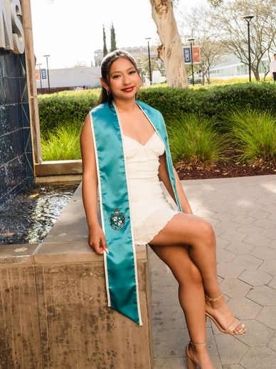 Young woman in white dress and teal graduation stole sitting outdoors.