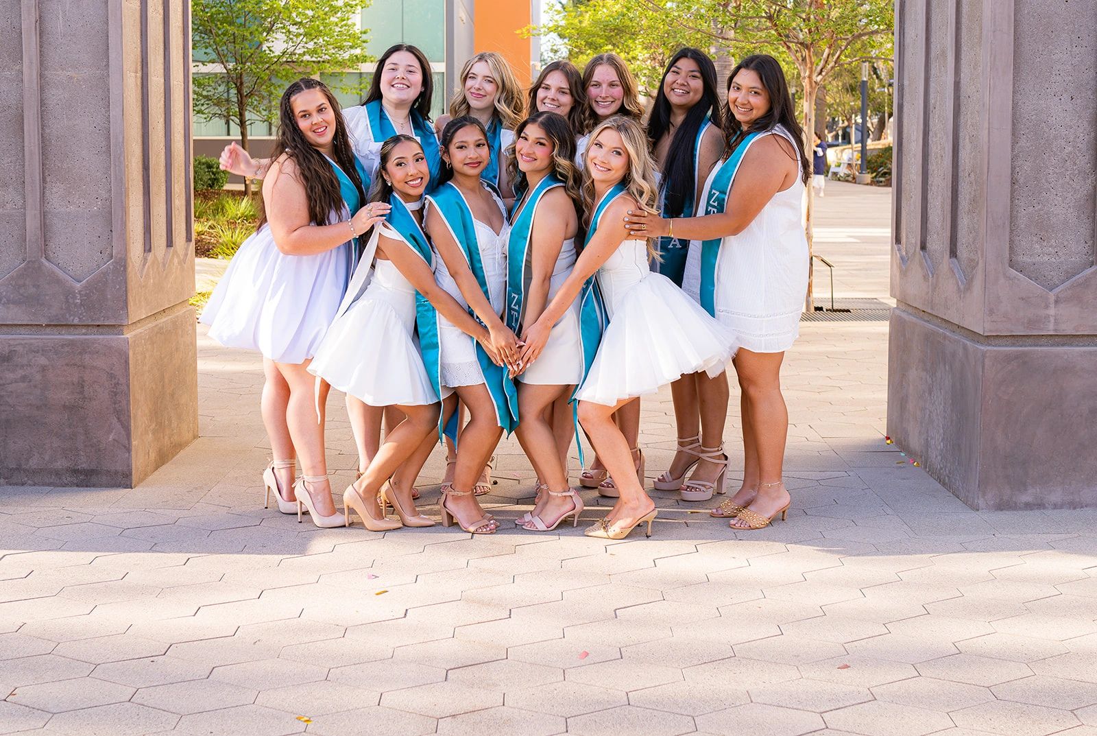 Group of women in white dresses and teal sashes posing outdoors.