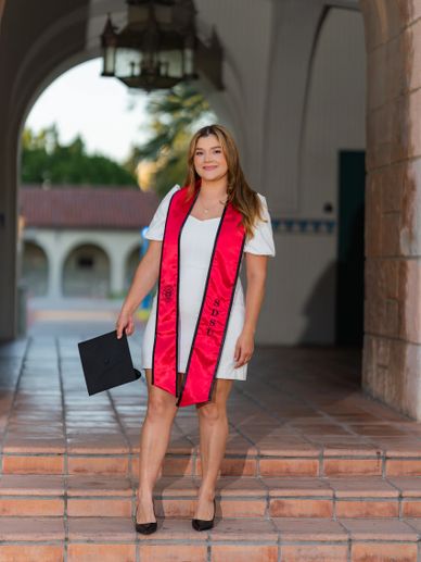 Young woman in white dress and red graduation stole holding cap on steps.
