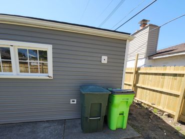 Two trash bins next to a gray house with a wooden fence.