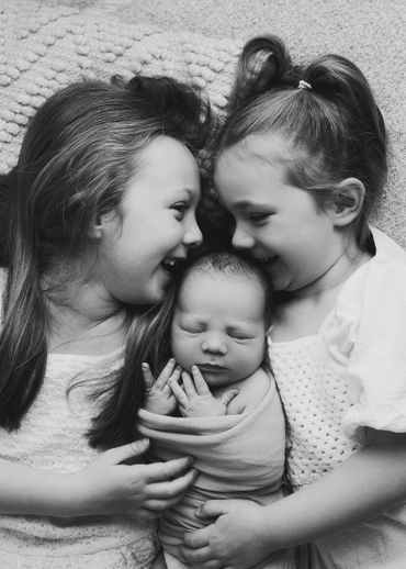 Two young girls lovingly embrace their newborn sibling in a tender black-and-white portrait.