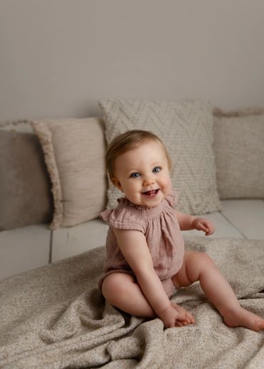 Smiling baby sitting on a cozy blanket with neutral-toned pillows.