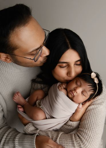 Parents lovingly hold and kiss their sleeping newborn baby wrapped in a soft blanket.