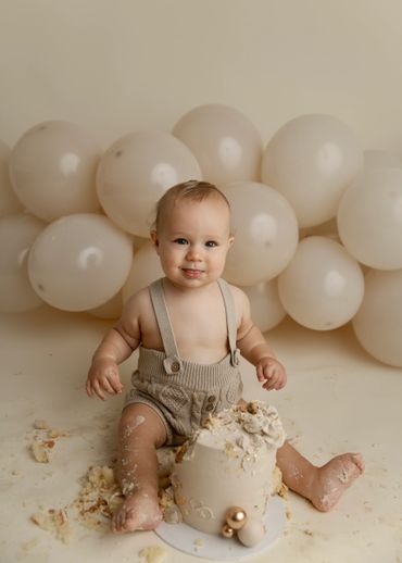 Baby in beige outfit enjoying a cake smash with cream balloons in the background.