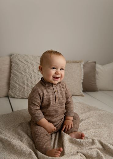 Smiling baby in cozy brown outfit sitting on a textured blanket indoors.