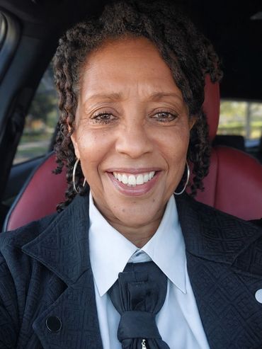 Smiling woman with braided hair in a car wearing a white shirt and black jacket.