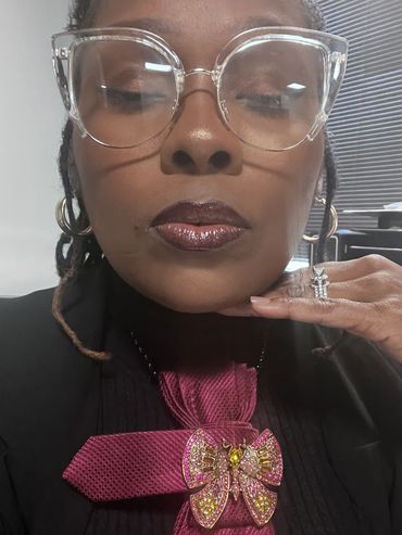 Woman with large clear glasses and a butterfly brooch on a magenta tie.