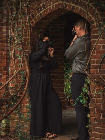 A man takes a photo of a woman posing by a brick archway covered in vines.