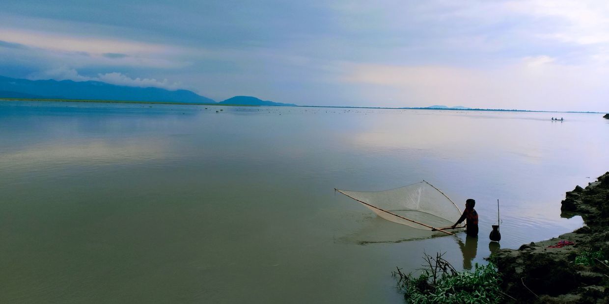 Person fishing with a net in calm water near a rocky shore.