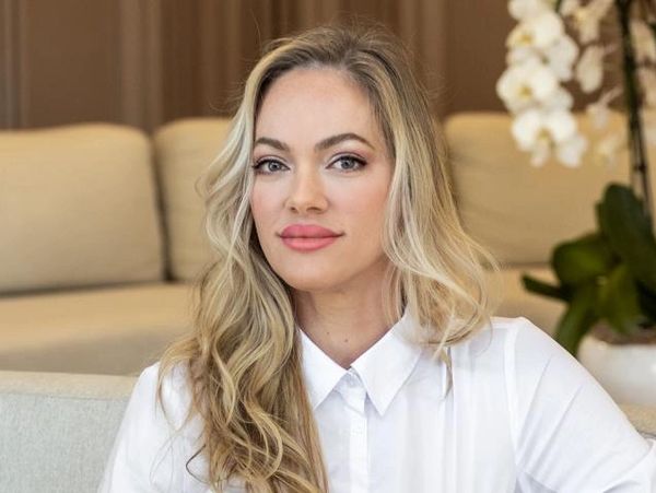 Confident woman with long blonde hair in a white shirt sits on a beige couch.