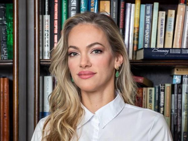 Confident woman with long blonde hair stands in front of a bookshelf filled with books.