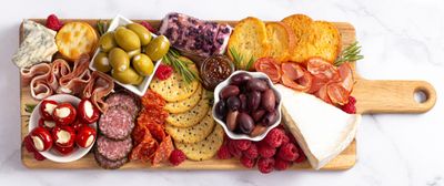 A colorful charcuterie board with cheeses, meats, olives, crackers, and raspberries.