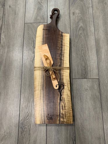 A handcrafted wooden serving board with utensils tied by a string on a wooden floor.