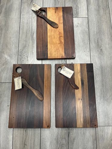 Three handcrafted wooden cutting boards with butter knives, displayed on a gray wood floor.