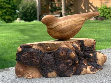 Wooden bird sculpture perched on a natural wood base outdoors.