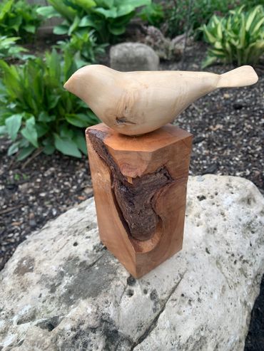 Wooden bird sculpture perched on a natural wood block outdoors.