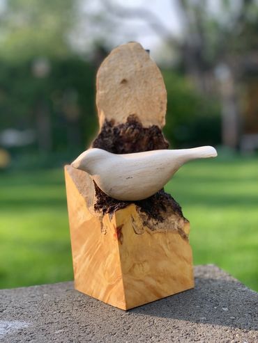 Wooden bird sculpture perched on a natural wood base outdoors.