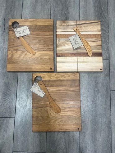 Three wooden cheese boards with spreader knives on a gray floor.